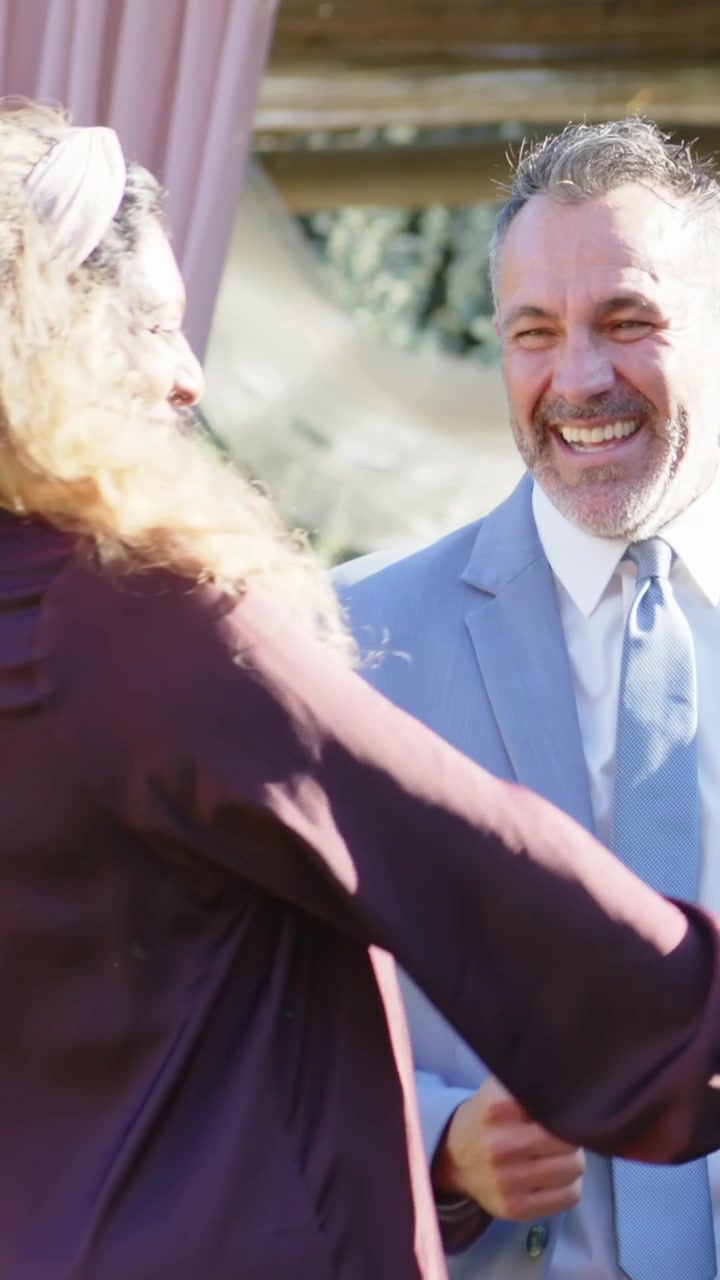 Video verticale: Donna sorridente con top color prugna che saluta mentre un uomo si avvicina da destra ridendo vicino a un pergolato.