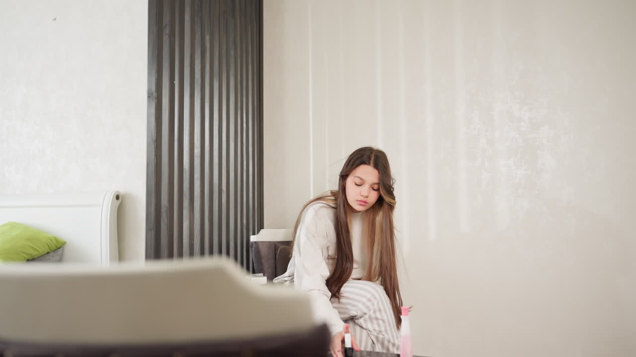 Beautiful lady walks past bed beside divider, moves to seat, sets nail polish on table, adjusts long hair, picks polish again, studies bottle, soft indoor light, modern room with subtle wall pattern