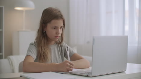 teen girl is learning distantly writing in exercise book and looking at display of notebook staying home