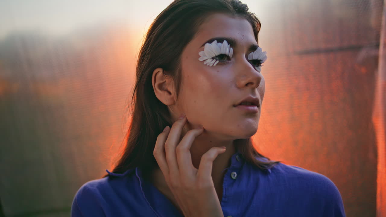 mujer original tocando el cabello posando con pestañas de pétalos en el resplandor del atardecer en primer plano.