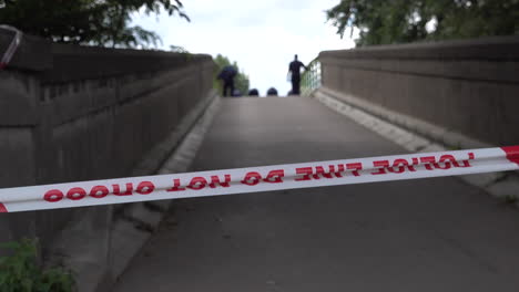 a london metropolitan police forensics team working on their hands and knees to investigate and search a bridge behind red police cordon tape, following a gun and knife crime incident