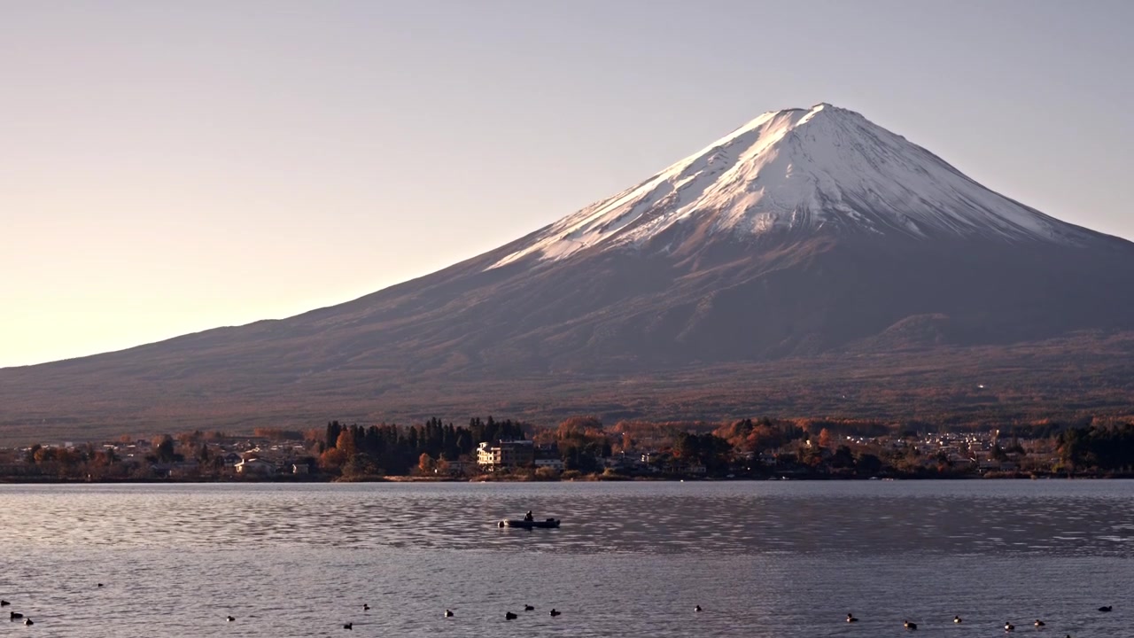 La telecamera cinematografica si inclina verso l'alto dalle calme acque del lago Kawaguchiko per rivelare l'iconica cima innevata del Monte Fuji contro un cielo limpido durante l'autunno in Giappone