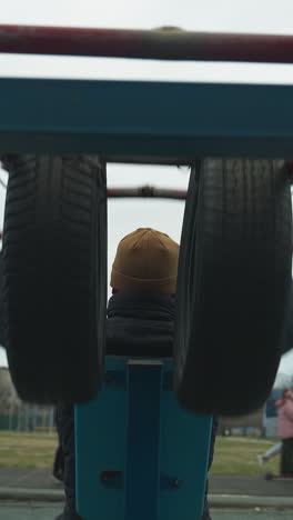 back view of a coach doing pull-ups on workout equipment while two boys stand in the background, with someone in pink riding an electric bike on a track nearby
