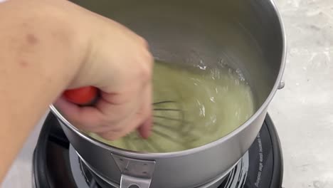 slow motion close up shot of a professional pastry chef hand mixing and whisking cooked egg white with steaming hot water on electric cooktop in a commercial bakery setting