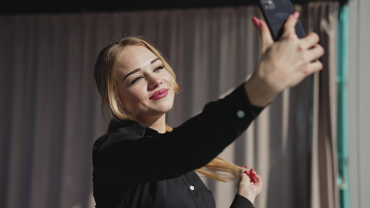 Beautiful red-haired woman taking selfie near the window. Attractive lady lit by the sun coming from panoramic window.