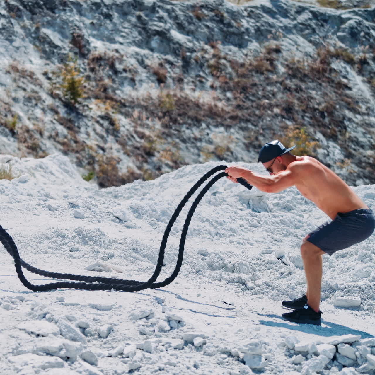 Side view on bodybuilder training with battling ropes outdoors. Handsome athlete quickly moves his hands with cables on the mountain background.