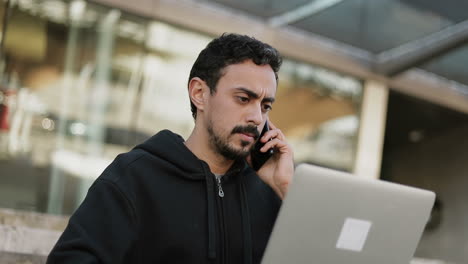 Concentrated-young-Arabic-man-with-dark-curly-hair-and-beard-in-black-hoodie-sitting-on-stairs-outside