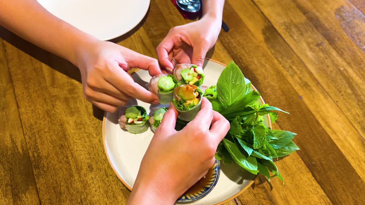 Hands assemble Vietnamese spring rolls with fresh herbs and dipping sauce on a wooden table under bright lighting