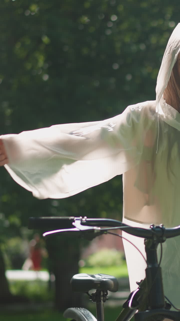 반투명한 레인코트를 입은 젊은 여성은 팔을 펴고 자전거 에 기게 서서 새의 날개처럼 손을 우아하게 흔들고, 빛은 잔잔한 초록색을 통해 뒷면에 건물을 필터링합니다.