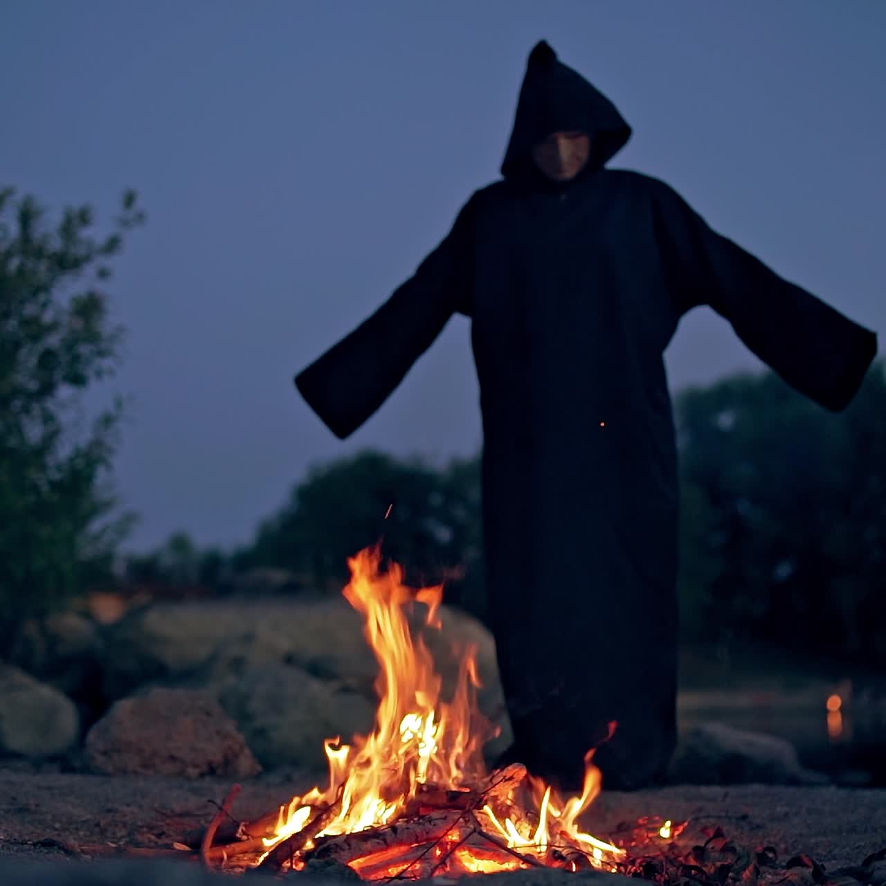 Spooky evening among nature. Dark nightmare by the the fire flame. Black wizard in mantle waving arms by the sides at the fire. Slow motion. Halloween.