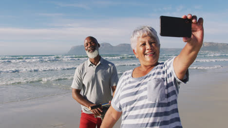 smiling senior african american couple walking with bicycles and taking a selfie at the beach