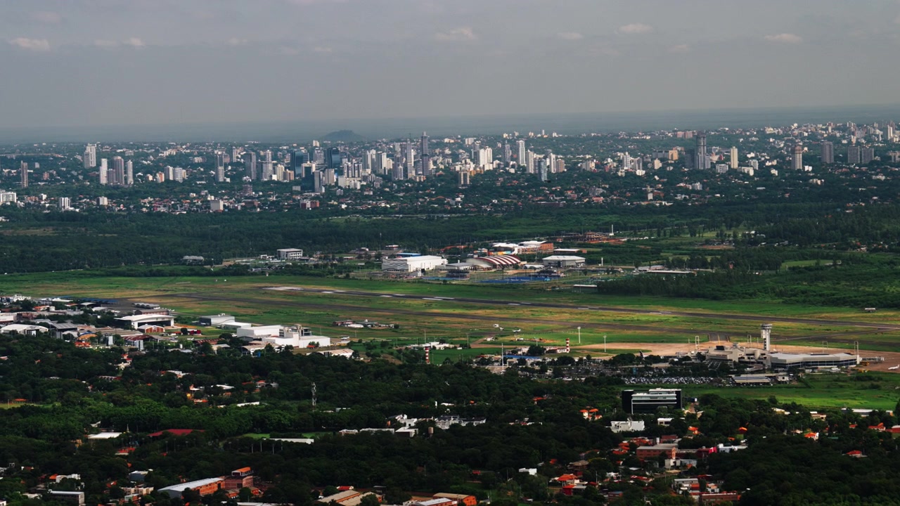 Letecký pohled na Asunción, Paraguay, ukazující panorama města, letištní dráhu a okolní městskou krajinu. Záběr z okna letadla, zdůrazňující infrastrukturu, cestování a rozvoj jihoamerického města.