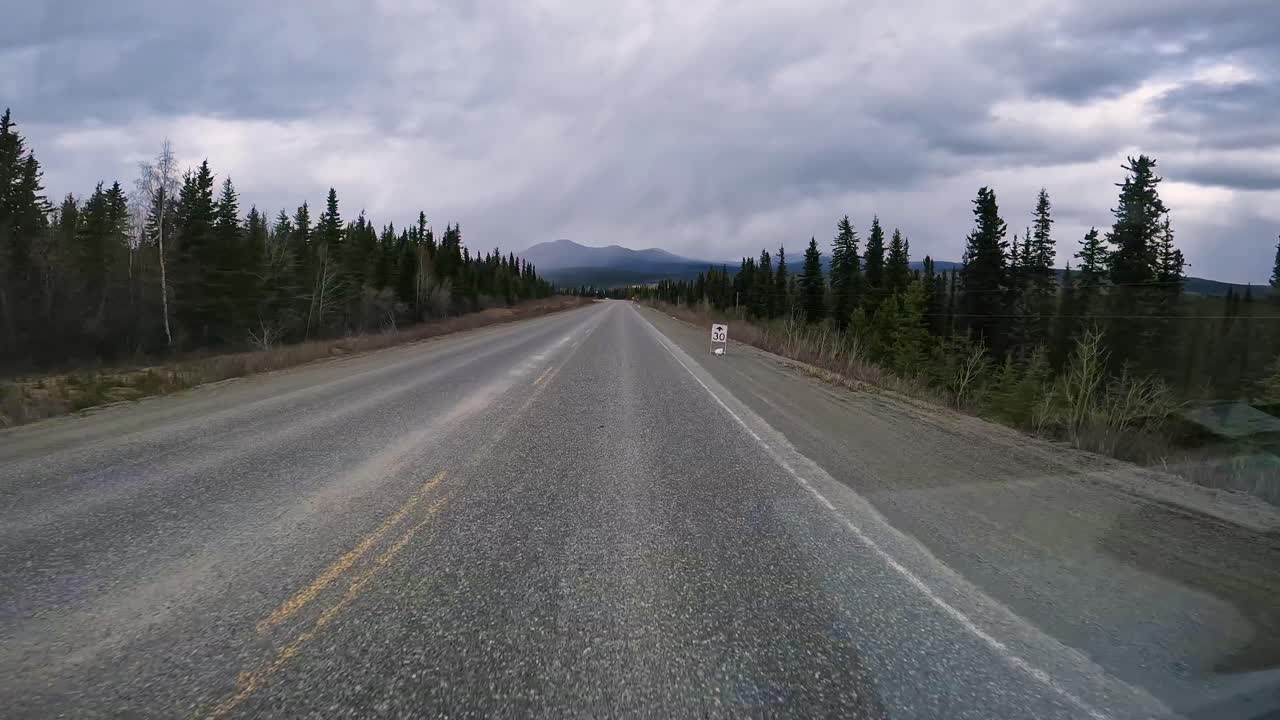 POV - driving on the Alaska Highway in the Yukon Territory; driving thru boreal forests with mountain peaks and Teslin Lake visible in the distance; concepts of wilderness, adventure, and travel