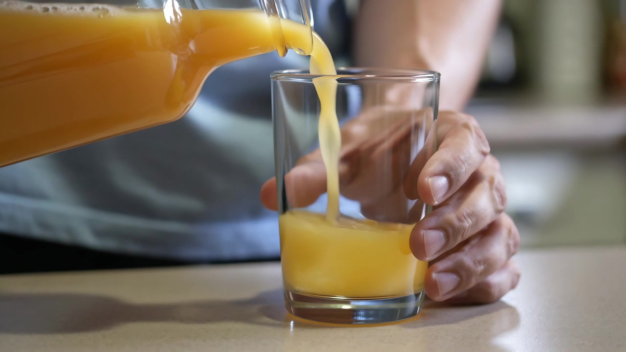 Manos vertiendo zumo de naranja, inclinando una jarra transparente y llenando un vaso en la encimera de la cocina
