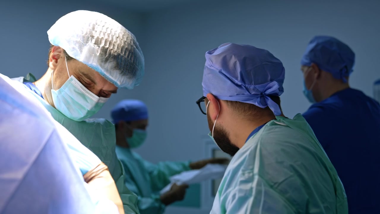 Due chirurghi uomini che conversano in sala operatoria. Il medico barbuto si rivolge agli assistenti chiedendo i materiali necessari.