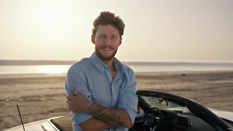 Portrait:-A-bearded-man-with-curly-hair-wearing-a-blue-shirt-is-crosses-his-arms-over-his-chest-and-shows-his-tattoos,-standing-next-to-a-white-convertible-against-the-yellow-sky-on-the-beach