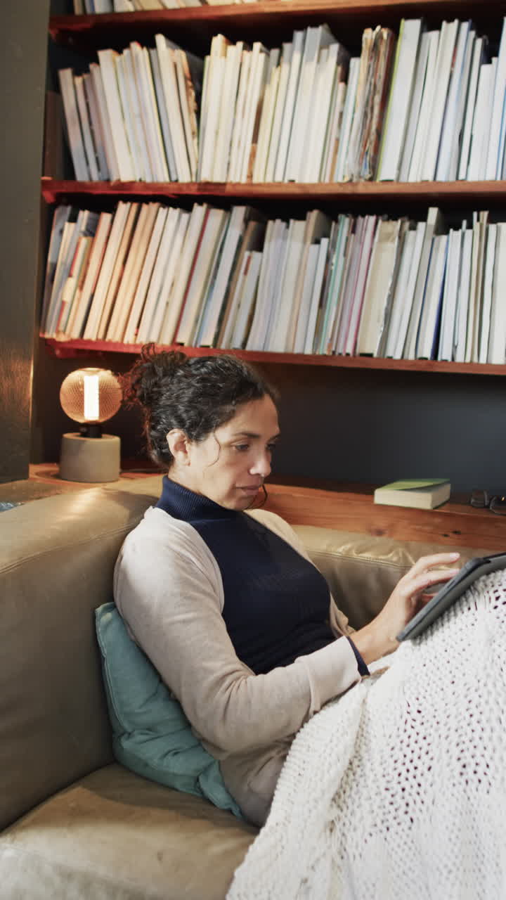 Vertical video of happy biracial woman lying on sofa and using tablet at home, slow motion