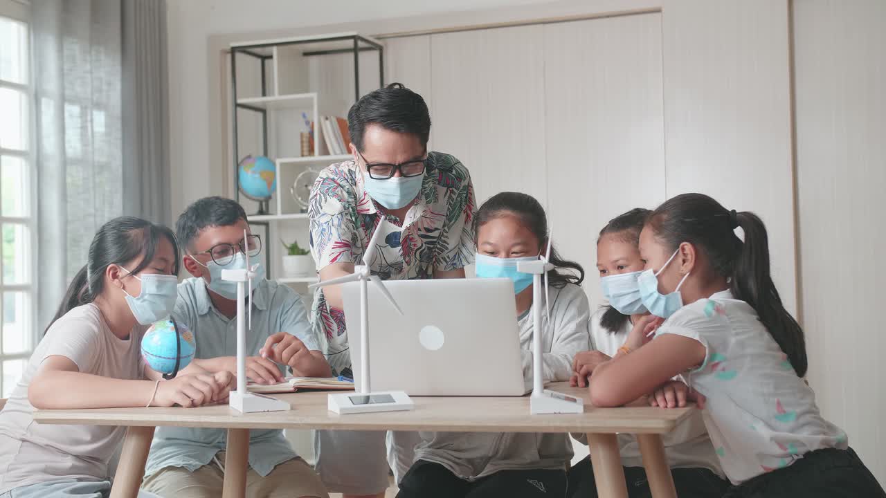 Middle School Classroom, Wearing Facemasks, Enthusiastic Asian Teacher Explains To Young Children How Wind Turbines Work. Kids Learning About Eco-Friendly Forms Of Renewable Energy