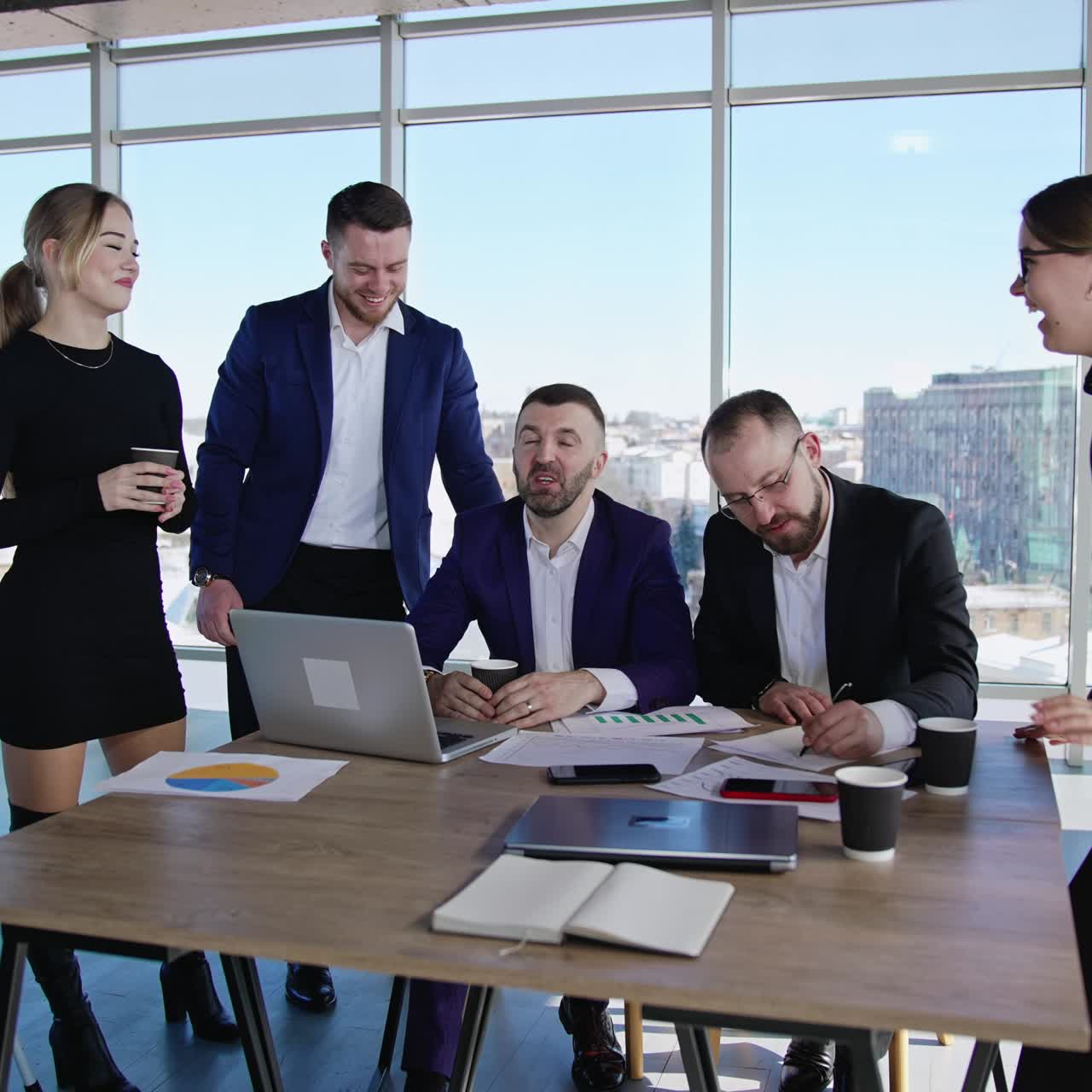 Teamwork and collaboration between colleagues. Male and female coworkers sit and stand around the table in the office. Cityscape at the background