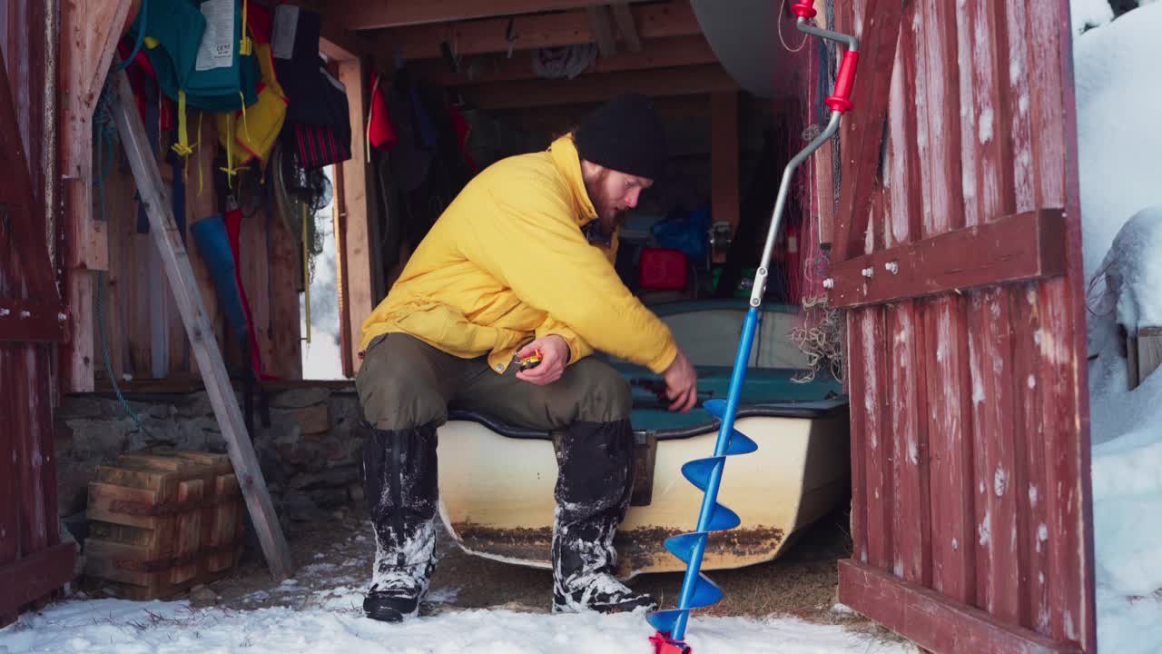 hombre en almacenamiento de madera se sienta en un bote mientras prepara equipo para pescar en hielo en invierno