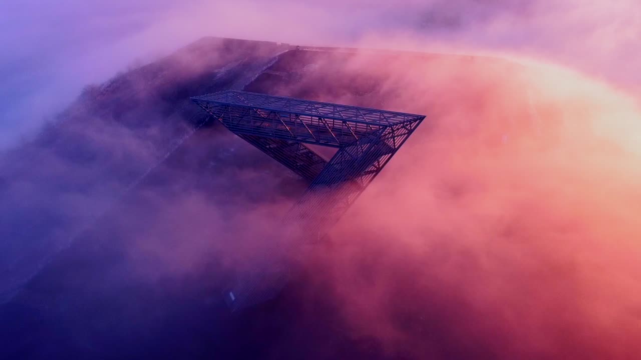 Monument on mountain in early morning light with sun shining from one side and clouds passing over the top, drone shot