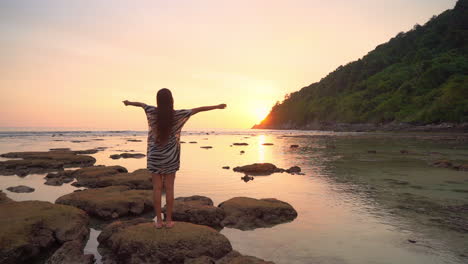 a young woman in a zebra print coverup stands on a large rock in the lagoon greets the rising sun