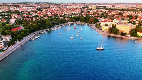 flying backwards over zadar’s coast, revealing a winding coastal road, a calm cove with anchored boats, deep blue sea, and soft sunset glow with the city fading in the distance.