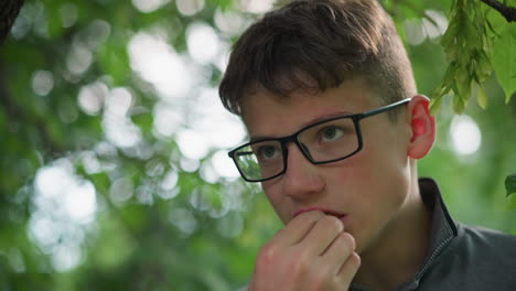 close-up of young boy wearing glasses and grey top eating a snack calmly under a tree, he appears deep in thought, surrounded by lush green leaves in a tranquil outdoor environment