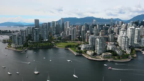 aerial view showing many boats on falls creek river and gigantic skyscraper tower of vancouver city in background - mountain range silhouette in backdrop - rising drone flight