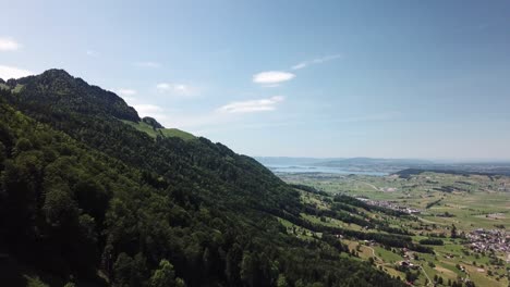 Drone-rises-up-on-a-green-forest-on-a-mountain-next-to-a-valley-with-some-villages-near-the-lake-of-Zurich-in-Switzerland-on-a-warm-summer-day