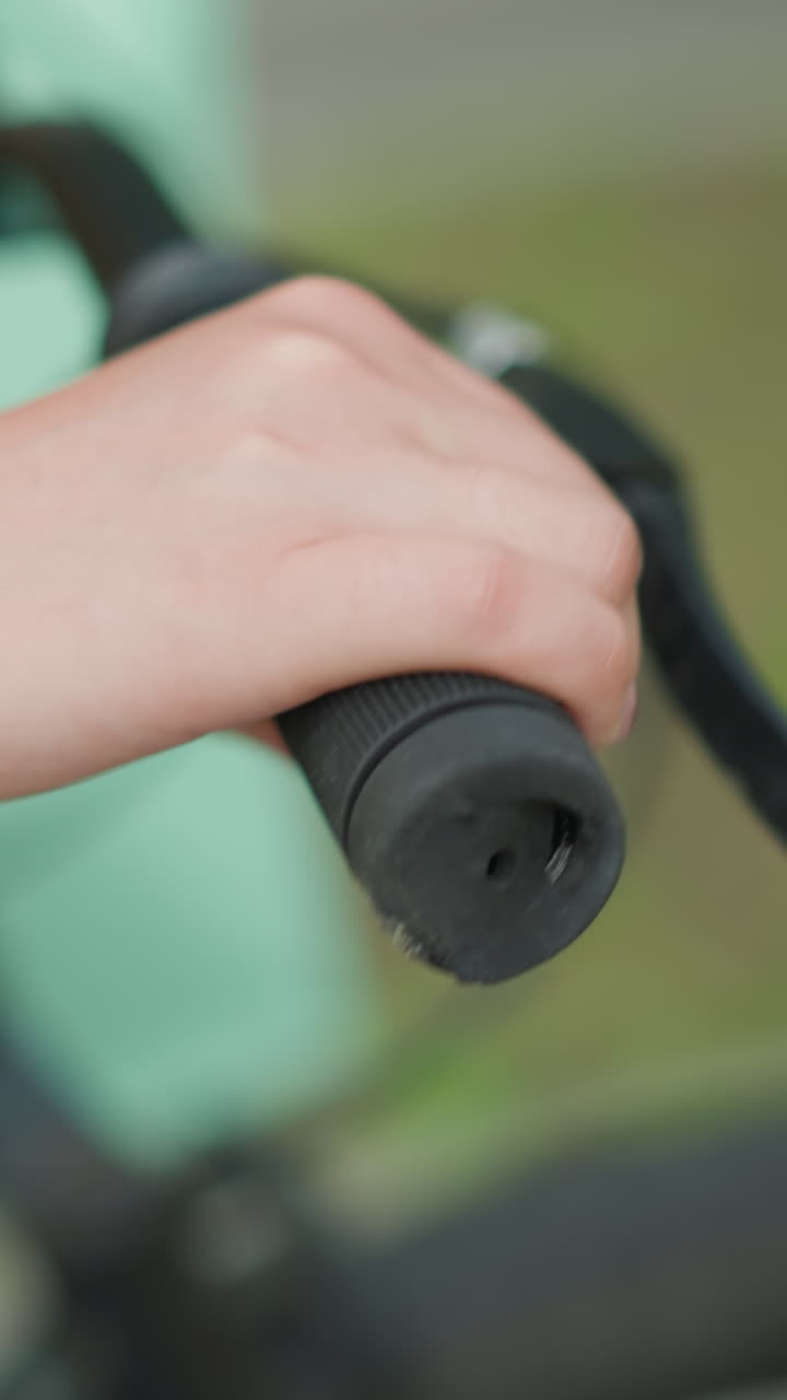 primer plano de una persona agarrando el manillar de una bicicleta, acompañada de una bolsa de color menta, mientras camina por un camino en un parque sereno con vegetación borrosa en el fondo