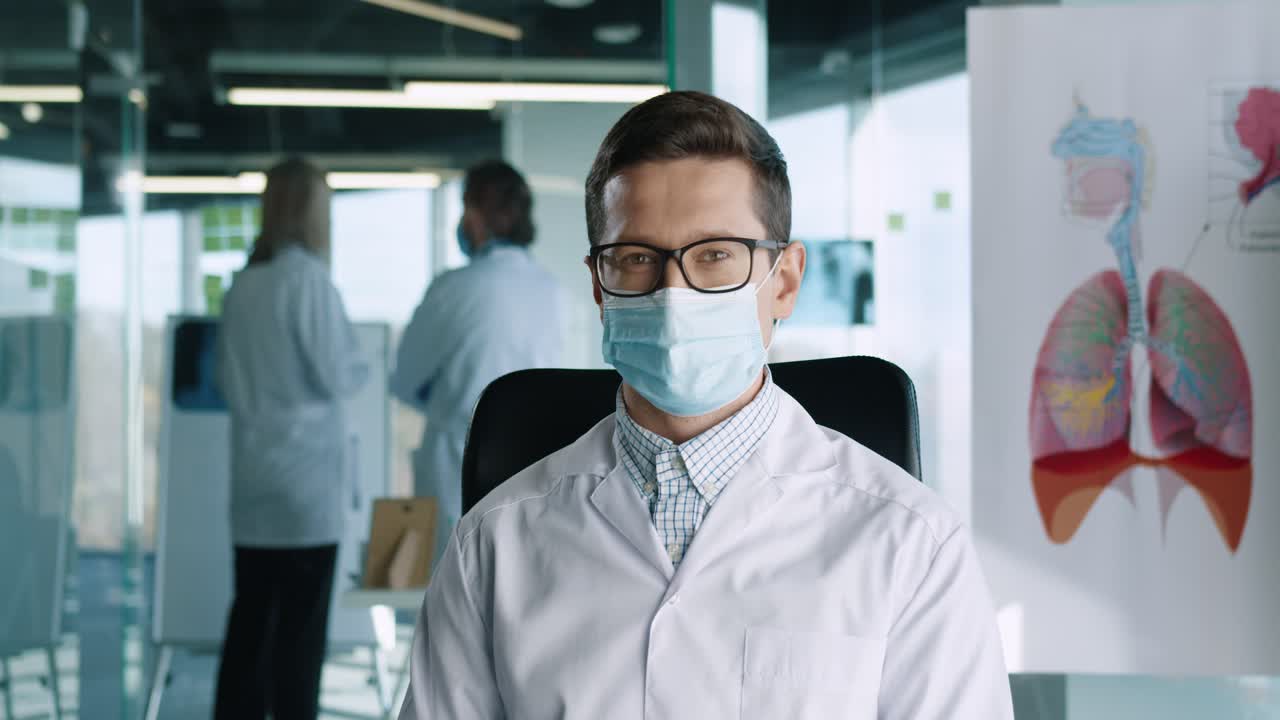 Close-up view of portrait of handsome young Caucasian adult male doctor wearing medical mask sitting in clinic at workplace looking at camera and smiling