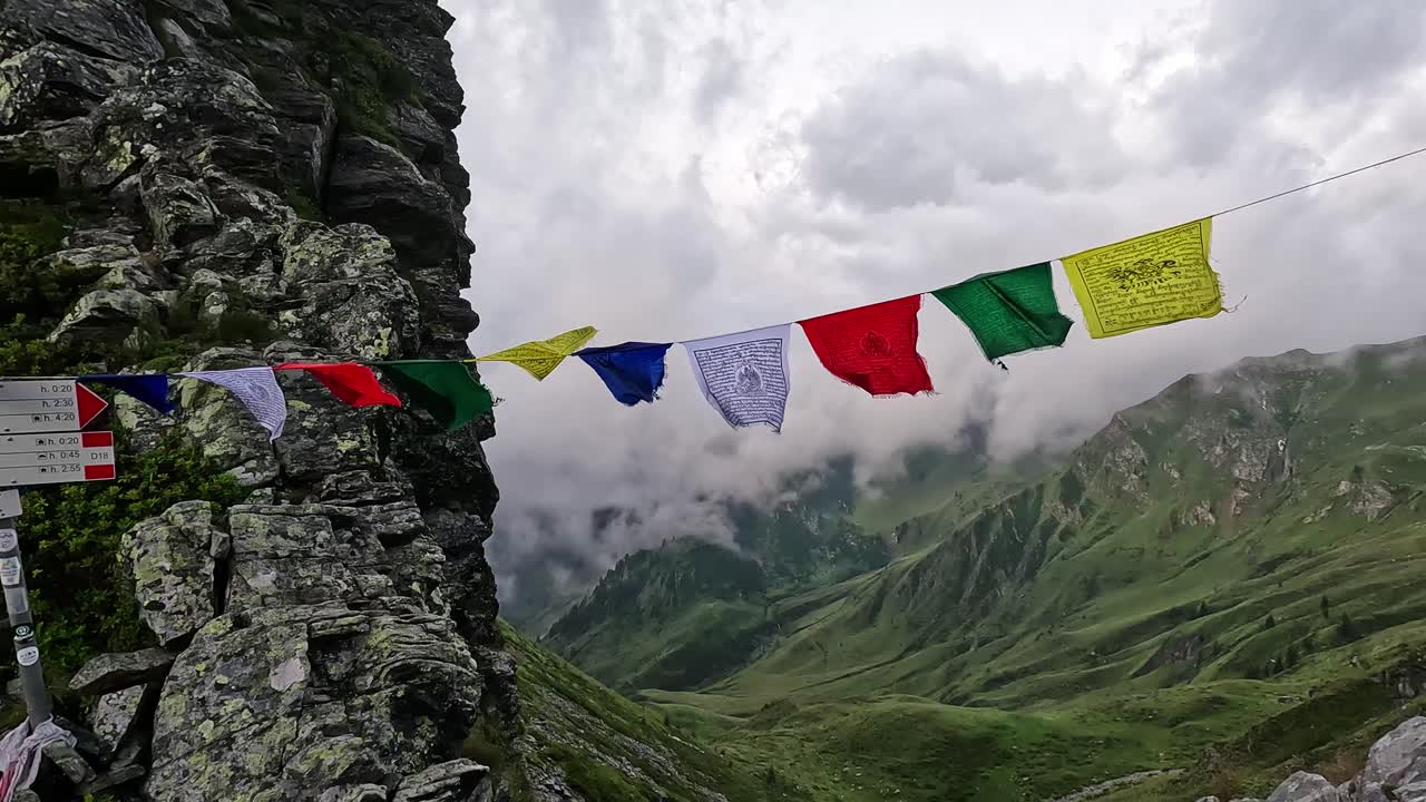 Vibrant Tibetan prayer flags sway in the wind on a rocky outcrop high above a lush, cloud-covered valley in the Lepontine Alps, Piedmont, Italy, creating a serene and spiritual alpine scene