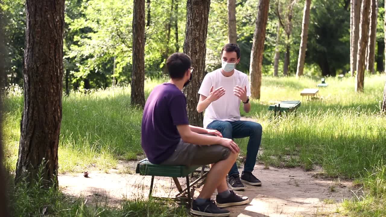 Two friends in protective medical masks talking in the park while resting on the bench by keeping their social distance from each other. Sunny day. Corona Virus idea