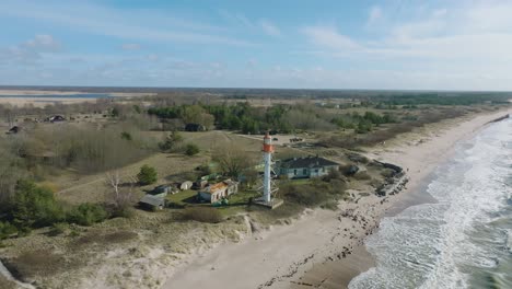 Aerial-establishing-view-of-white-colored-Pape-lighthouse,-Baltic-sea-coastline,-Latvia,-white-sand-beach,-large-waves-crashing,-sunny-day-with-clouds,-wide-revealing-drone-shot-moving-backward