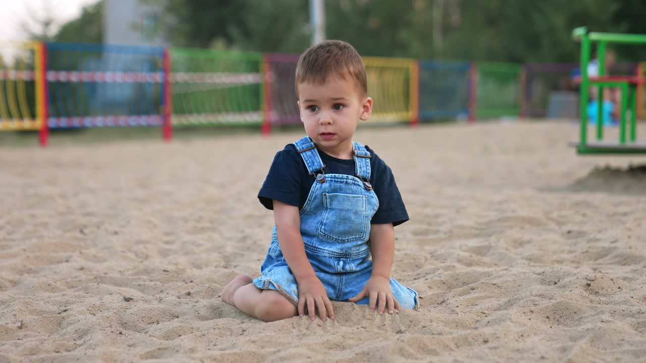 Cute baby boy in jeans romper sits in the sand outdoors. Kid deeps his hands into sand looking attentively away.