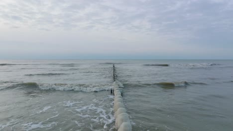 Aerial-establishing-view-of-an-old-wooden-pier-at-the-Baltic-sea-coastline,-overcast-winter-day,-white-sand-beach-covered-in-snow,-ice-on-wood-poles,-calm-seashore,-wide-drone-shot-moving-back-low