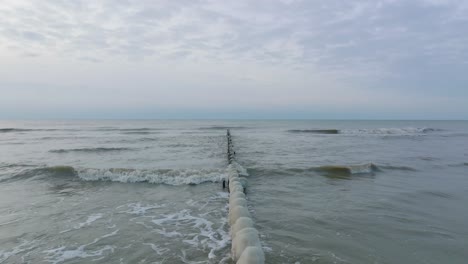 vista aérea de un antiguo muelle de madera en la costa del mar báltico, día de invierno nublado, playa de arena blanca cubierta de nieve, hielo en postes de madera, costa tranquila, amplio tiro de drones retrocediendo bajo