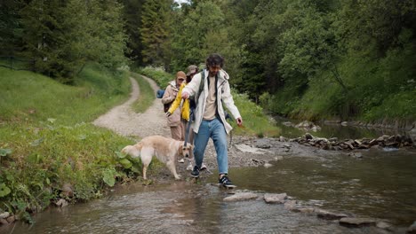 A-guy-and-two-girls-in-hiking-clothes,-together-with-their-dog,-are-trying-to-cross-a-mountain-river-along-a-special-path-made-of-stones