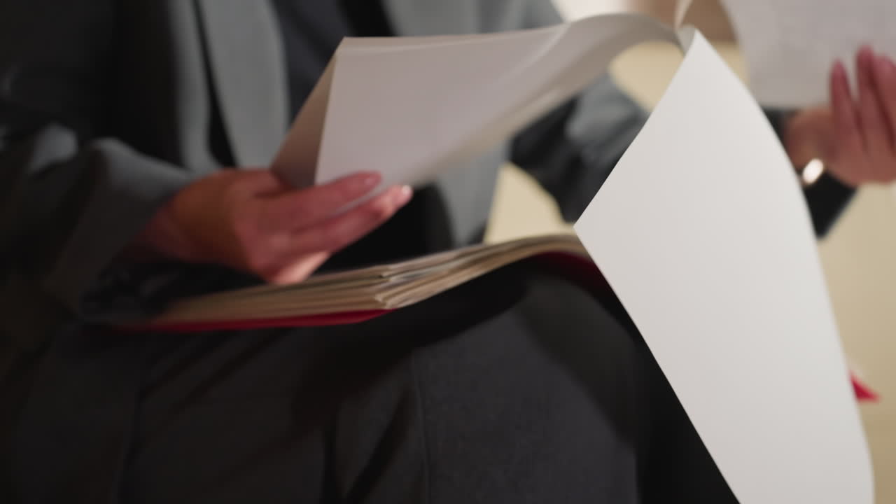 Close-up of businesswoman in gray suit flipping through large stack of documents in red folder while sitting in office, showcasing detail-oriented work, organization, and hands-on paper handling process