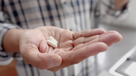 midsection of senior biracial man at home holding two pills in palm of hand, slow motion