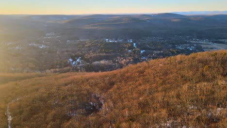 Drone-flying-over-sun-dappled-hills-and-into-a-valley-lightly-covered-in-snow,-with-mountains-in-the-distance-during-winter