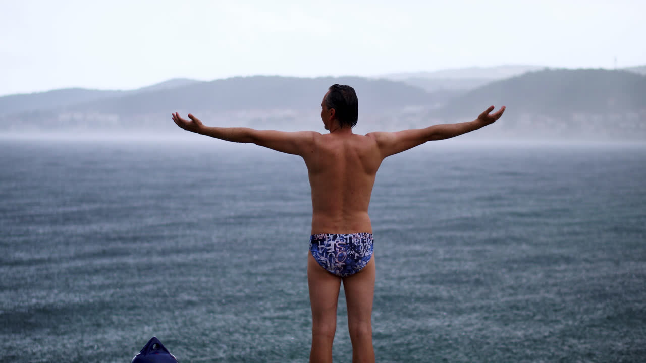vista trasera de un hombre en traje de baño de pie con los brazos extendidos en la playa