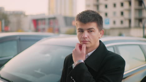 business mogul standing near luxury car lost in thought with hand on jaw, background featuring modern office building and parked cars in city setting