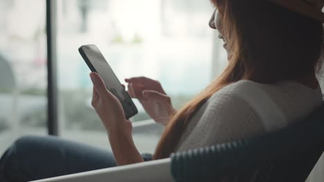 close up happy young woman sit on a armchair near the window at hotel while use mobile phone communication, smile female holding smartphone with internet and social media.