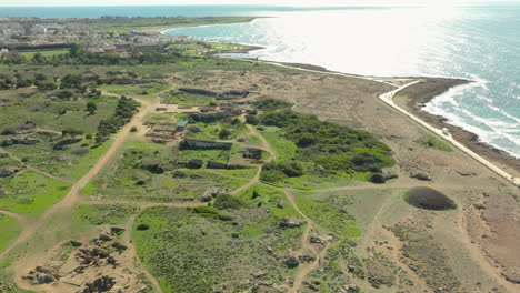 A-sweeping-aerial-view-of-the-Tombs-of-the-Kings-archaeological-site-in-Paphos,-Cyprus,-with-ancient-stone-structures-scattered-across-a-grassy-landscape-that-leads-to-a-rugged-coastline-Sea