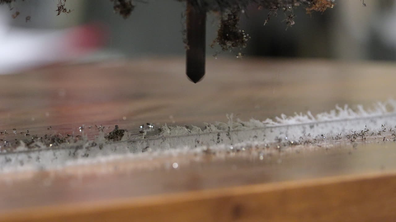 Macro di una punta per fresatrice CNC e un accessorio a spazzola che intagliano una scanalatura precisa su una tavola di legno, spargendo trucioli in una falegnameria professionale.