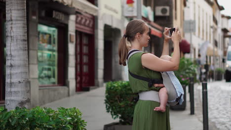 accompanied by her baby in a kangaroo backpack, a young woman captures memories on her mobile phone during the journey. she walks and periodically checks the screen