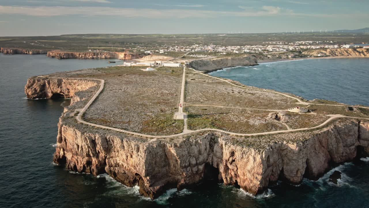 Aerial drone shot moving forward above the ocean, approaching the iconic cliffs, lighthouse, and fortress at the tip of Sagres, Portugal