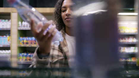 Mujer-Negra-Haciendo-Compras,-Tomando-Una-Botella-De-Agua-Del-Estante---Imágenes-Del-Interior